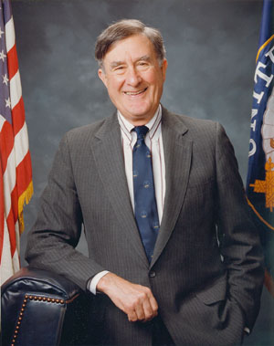 Portrait of Senator Chafee in a office setting with a grey pinstripe suit and flanked by the U.S. flag to his left and the Rhode Island state flag to his right